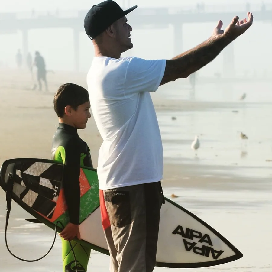 Kolby standing on the beach with a young boy holding a surfboard